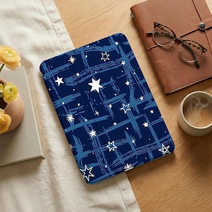 May include: A navy blue tablet case with a white star pattern and blue brushstroke design. The case is on a wooden surface next to a leather-bound journal, glasses, and a cup of coffee. The case is designed to protect a tablet.