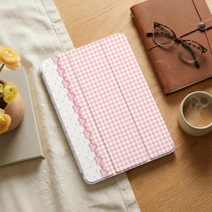Può includere: Una custodia per tablet a quadretti rosa e bianchi con un bordo decorativo di cuori rosa e pizzo bianco. La custodia è aperta, rivelando l'interno. La custodia è su una superficie di legno, con un diario rilegato in pelle, occhiali e una tazza di caffè nelle vicinanze.