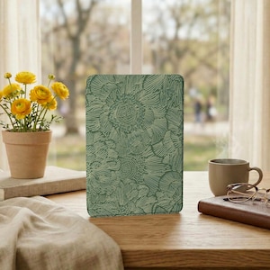 Puede incluir: Una funda verde para tableta con un estampado floral. La funda es rectangular y está sobre una mesa de madera. A la izquierda hay una pequeña planta en maceta con flores amarillas. Una taza de café y un libro también están sobre la mesa.