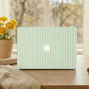 May include: A laptop with a light green and white striped design. The Apple logo is visible in the center. The laptop is on a wooden table, with a potted plant of yellow flowers to the left.