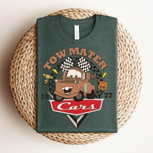 Puede incluir: Una camiseta verde oscuro con un gr&aacute;fico del personaje Tow Mater de la pel&iacute;cula Cars. El dise&ntilde;o incluye el texto Tow Mater, una bandera a cuadros, una se&ntilde;al de la Ruta 66, un trofeo y el logotipo de Cars con los n&uacute;meros 19 y 77 a los lados.