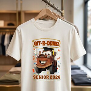 Op de afbeelding: Cr&egrave;mekleurig T-shirt met een cartoon sleepwagen met een afstudeerhoed. Het shirt heeft de tekst "GIT-R-DONE!" en "SENIOR 2026" met confetti en een geblokt vlagontwerp. Het shirt hangt aan een houten hanger.