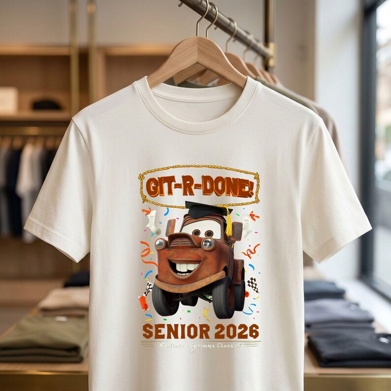 Op de afbeelding: Cr&egrave;mekleurig T-shirt met een cartoon sleepwagen met een afstudeerhoed. Het shirt heeft de tekst "GIT-R-DONE!" en "SENIOR 2026" met confetti en een geblokt vlagontwerp. Het shirt hangt aan een houten hanger.