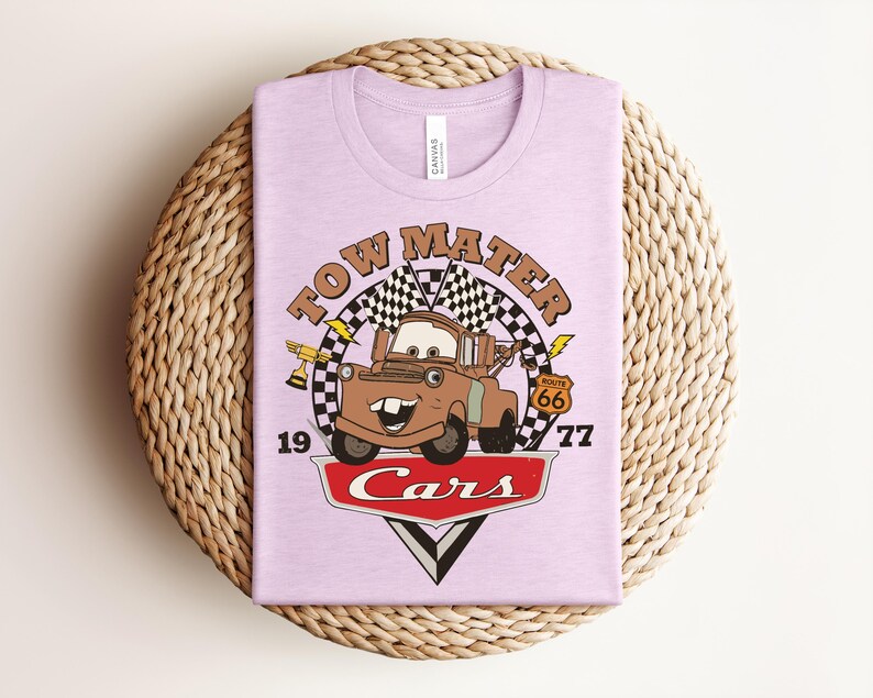 Puede incluir: Una camiseta de color morado claro con un gr&aacute;fico del personaje Tow Mater de la pel&iacute;cula Cars. El dise&ntilde;o incluye una bandera a cuadros, un trofeo dorado, una se&ntilde;al de la Ruta 66, el texto Tow Mater, 1977, y el logo de Cars en rojo y blanco.