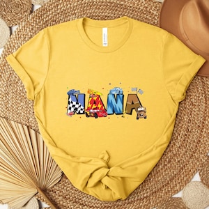 K&ouml;nnte beinhalten: Senfgelbes T-Shirt mit dem Wort "NANA" in farbenfrohen, cartoonartigen Buchstaben. Jeder Buchstabe ist mit einem anderen Auto-Thema gestaltet, einschlie&szlig;lich Rennwagen und Zielflaggen. Ein l&auml;ssiges Kleidungsst&uuml;ck.