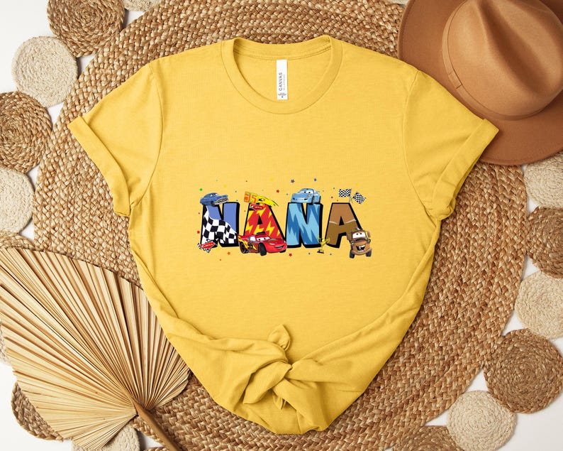 K&ouml;nnte beinhalten: Senfgelbes T-Shirt mit dem Wort "NANA" in farbenfrohen, cartoonartigen Buchstaben. Jeder Buchstabe ist mit einem anderen Auto-Thema gestaltet, einschlie&szlig;lich Rennwagen und Zielflaggen. Ein l&auml;ssiges Kleidungsst&uuml;ck.