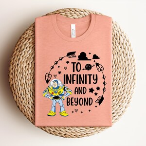 Peut inclure: Un t-shirt corail avec un motif de Buzz l'&Eacute;clair et l'inscription "To Infinity and Beyond" en noir. Le motif comprend des &eacute;toiles, des fl&egrave;ches et une plan&egrave;te. Le t-shirt est pli&eacute; sur un tapis circulaire tiss&eacute;.