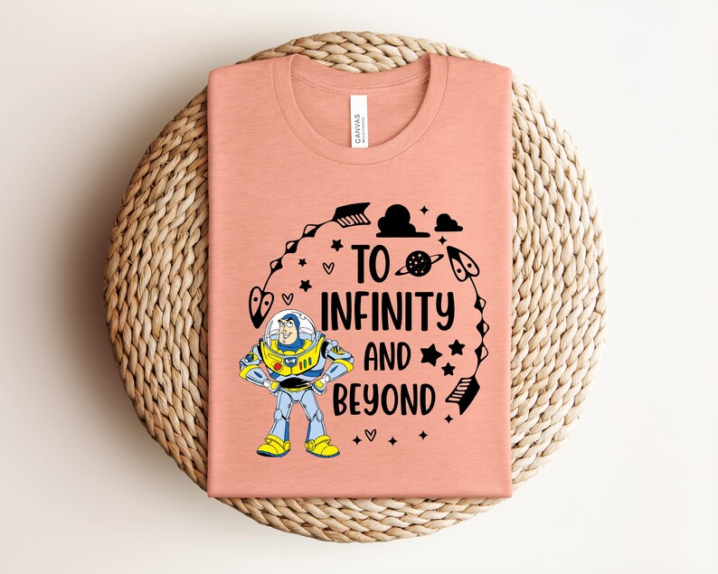 Peut inclure: Un t-shirt corail avec un motif de Buzz l'&Eacute;clair et l'inscription "To Infinity and Beyond" en noir. Le motif comprend des &eacute;toiles, des fl&egrave;ches et une plan&egrave;te. Le t-shirt est pli&eacute; sur un tapis circulaire tiss&eacute;.