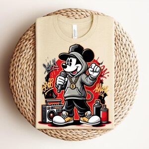 May include: Beige t-shirt featuring a cartoon Mickey Mouse in a hip-hop style. Mickey is wearing a gray hoodie, black pants, and white sneakers with yellow accents. He holds a microphone and is set against a red backdrop with graffiti art.
