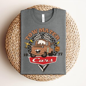 Puede incluir: Una camiseta gris jaspeado con un gr&aacute;fico del personaje Tow Mater de la pel&iacute;cula Cars. El dise&ntilde;o incluye el texto Tow Mater, Route 66 y Cars, junto con banderas a cuadros, un trofeo, rayos y los n&uacute;meros 19 y 77 impresos en la parte frontal.