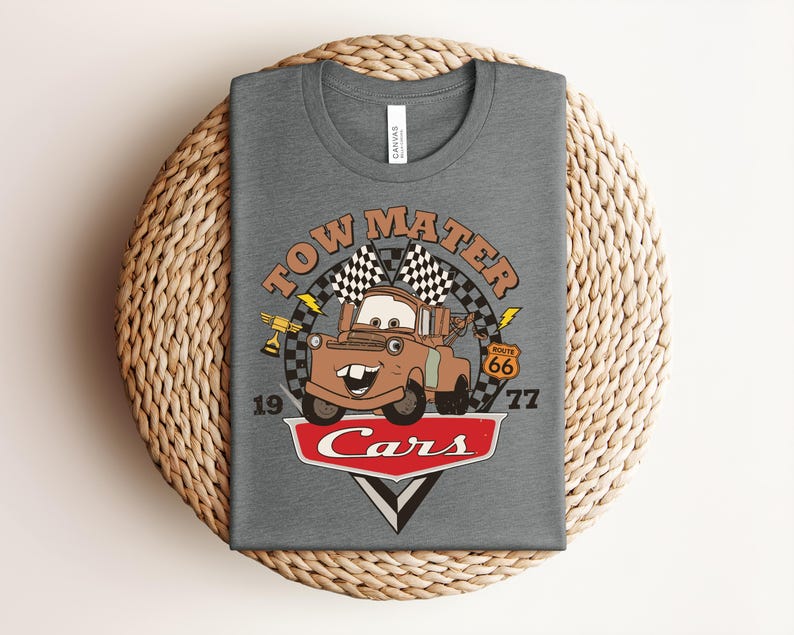 Puede incluir: Una camiseta gris jaspeado con un gr&aacute;fico del personaje Tow Mater de la pel&iacute;cula Cars. El dise&ntilde;o incluye el texto Tow Mater, Route 66 y Cars, junto con banderas a cuadros, un trofeo, rayos y los n&uacute;meros 19 y 77 impresos en la parte frontal.