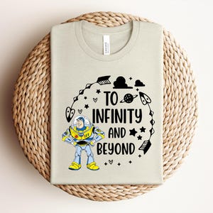 Peut inclure: Un t-shirt beige clair avec un dessin de Buzz l'&Eacute;clair et l'inscription "TO INFINITY AND BEYOND" en noir. Le motif comprend des &eacute;toiles, des plan&egrave;tes et une fl&egrave;che. Le t-shirt est pr&eacute;sent&eacute; sur une surface circulaire tiss&eacute;e.