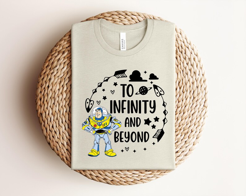 Peut inclure: Un t-shirt beige clair avec un dessin de Buzz l'&Eacute;clair et l'inscription "TO INFINITY AND BEYOND" en noir. Le motif comprend des &eacute;toiles, des plan&egrave;tes et une fl&egrave;che. Le t-shirt est pr&eacute;sent&eacute; sur une surface circulaire tiss&eacute;e.