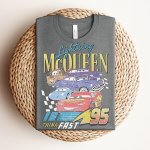 Puede incluir: Camiseta gris oscuro con un dise&ntilde;o de Rayo McQueen y otros personajes de Cars. El dise&ntilde;o incluye el texto "Lightning McQueen" en azul y amarillo, y "Think Fast" con el n&uacute;mero 95.