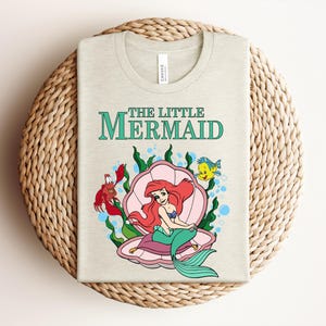 May include: A light beige t-shirt featuring a graphic print of Ariel from The Little Mermaid sitting inside a pink seashell. The design includes Sebastian the crab, Flounder the fish, green seaweed, and blue bubbles with the text The Little Mermaid.