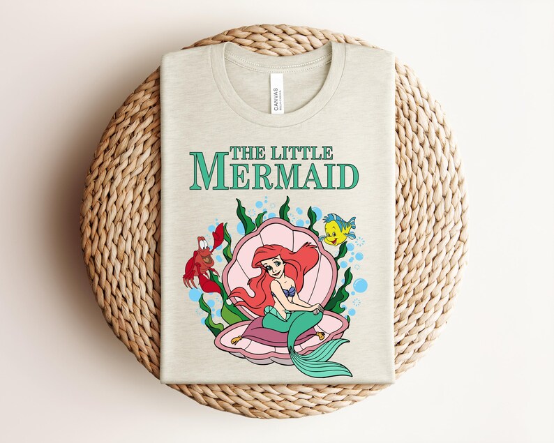 May include: A light beige t-shirt featuring a graphic print of Ariel from The Little Mermaid sitting inside a pink seashell. The design includes Sebastian the crab, Flounder the fish, green seaweed, and blue bubbles with the text The Little Mermaid.