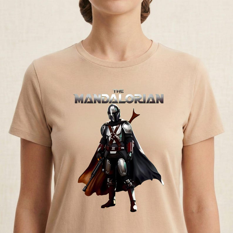 Op de afbeelding: Beige T-shirt met een afbeelding van The Mandalorian. De afbeelding toont het personage in volledige bepantsering, met een zwarte cape en een geweer. De tekst "THE MANDALORIAN" staat in een metallic lettertype boven het personage.