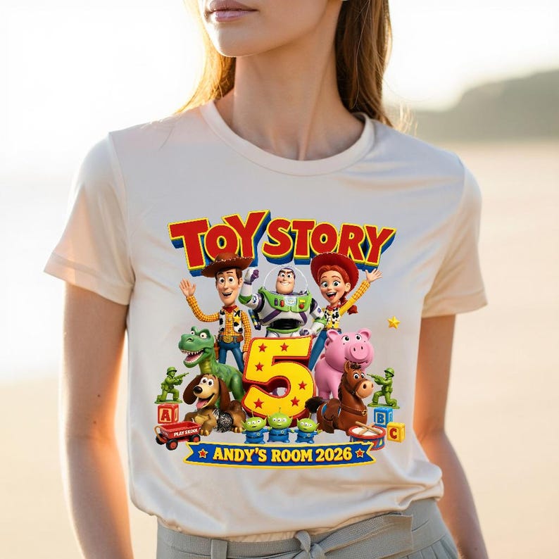 May include: A cream-colored t-shirt featuring a colorful Toy Story graphic with characters Woody, Buzz Lightyear, Jessie, Rex, Slinky Dog, Hamm, Bullseye, and green army men. The design includes a large yellow number 5 and text reading Toy Story Andy's Room 2026.