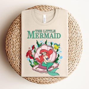 May include: A beige t-shirt featuring a graphic of The Little Mermaid with Ariel sitting in a pink seashell, accompanied by a red crab and a yellow fish, surrounded by green seaweed and blue bubbles, with the text The Little Mermaid printed above.