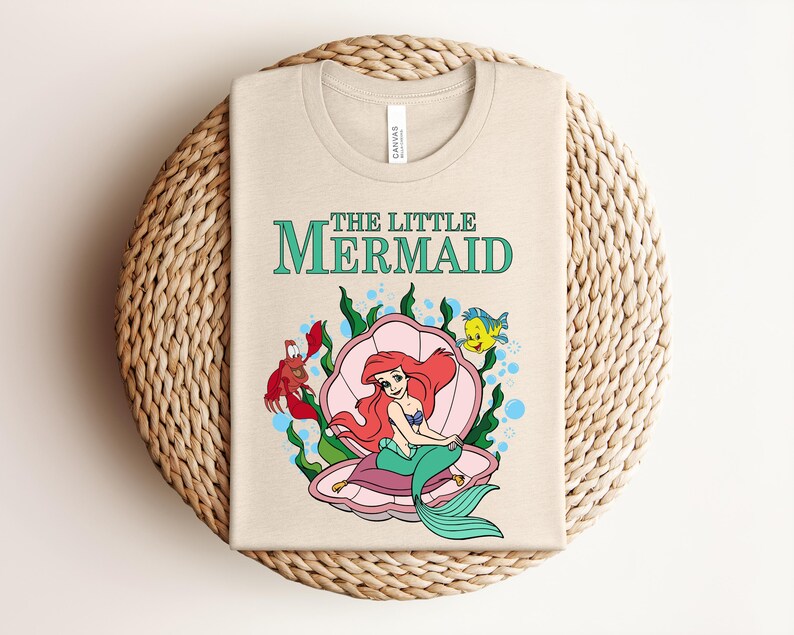 May include: A beige t-shirt featuring a graphic of The Little Mermaid with Ariel sitting in a pink seashell, accompanied by a red crab and a yellow fish, surrounded by green seaweed and blue bubbles, with the text The Little Mermaid printed above.