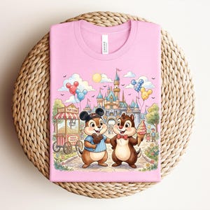 May include: Pink t-shirt featuring a whimsical illustration of Chip and Dale at a theme park. The design includes a popcorn stand, balloons, a castle, and the text "Magic Treats". The shirt is made of soft material.