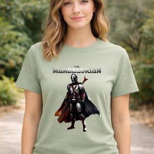 Op de afbeelding: Saliegroen T-shirt met de tekst "THE MANDALORIAN" en een afbeelding van de Mandalorian. De afbeelding toont het personage in harnas, cape en wapen. Het T-shirt heeft een ronde hals.