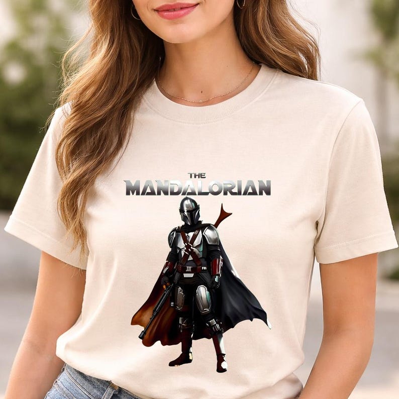 Op de afbeelding: Beige T-shirt met een afbeelding van The Mandalorian. Het ontwerp bevat de tekst "THE MANDALORIAN" boven een illustratie van het personage in volledige uitrusting, cape en wapen. Het T-shirt heeft een ronde hals en korte mouwen.