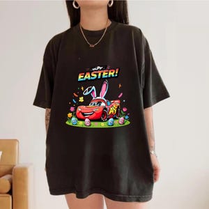 Puede incluir: Camiseta negra con un gr&aacute;fico colorido de un coche de carreras rojo con orejas de conejo, rodeado de huevos de Pascua y flores. El texto "HAPPY EASTER!" est&aacute; encima del coche. Un dise&ntilde;o divertido y festivo para la Pascua.