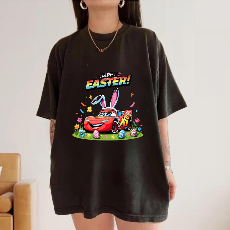 Puede incluir: Camiseta negra con un gr&aacute;fico colorido de un coche de carreras rojo con orejas de conejo, rodeado de huevos de Pascua y flores. El texto "HAPPY EASTER!" est&aacute; encima del coche. Un dise&ntilde;o divertido y festivo para la Pascua.