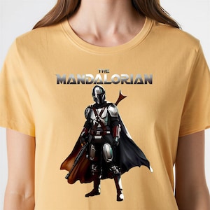 Op de afbeelding: Mosterdgeel T-shirt met een afbeelding van The Mandalorian. De afbeelding toont het personage in volledige bepantsering met een zwarte cape en een blastergeweer. De tekst "THE MANDALORIAN" staat in zilver boven het personage.