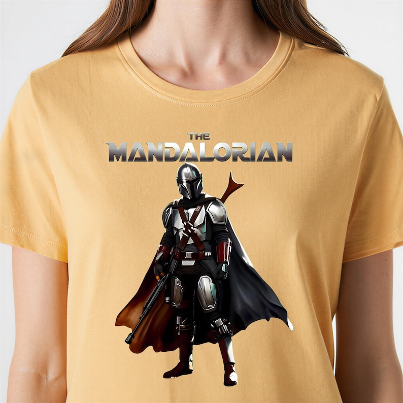 Op de afbeelding: Mosterdgeel T-shirt met een afbeelding van The Mandalorian. De afbeelding toont het personage in volledige bepantsering met een zwarte cape en een blastergeweer. De tekst "THE MANDALORIAN" staat in zilver boven het personage.