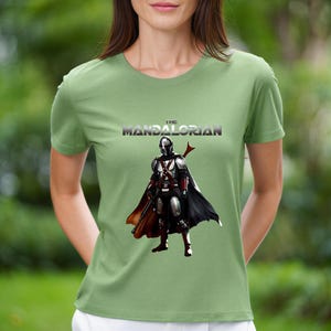 Op de afbeelding: Saliegroen T-shirt met de tekst "THE MANDALORIAN" boven een afbeelding van het Mandalorian-personage in harnas en cape. De afbeelding is in zwart, zilver, rood en bruin.