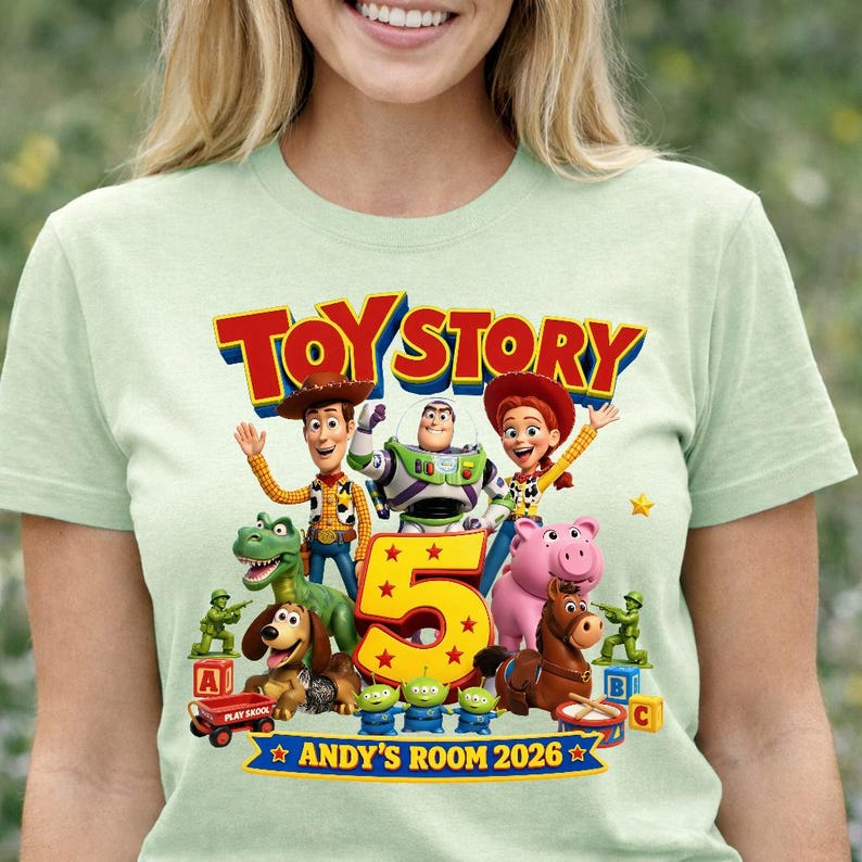 May include: A light green t-shirt featuring a colorful Toy Story graphic with characters Woody, Buzz Lightyear, Jessie, Rex, Slinky Dog, Hamm, Bullseye, and green army men surrounding a large yellow number 5 and the text Toy Story 2026.