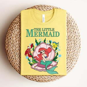 May include: A yellow t-shirt featuring a graphic of The Little Mermaid with Ariel sitting in a pink seashell, accompanied by a red crab and a yellow fish. The text THE LITTLE MERMAID is printed in teal above the illustration, which includes green seaweed.