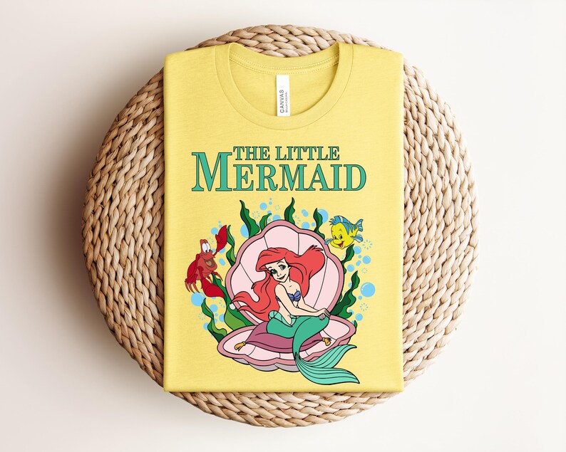 May include: A yellow t-shirt featuring a graphic of The Little Mermaid with Ariel sitting in a pink seashell, accompanied by a red crab and a yellow fish. The text THE LITTLE MERMAID is printed in teal above the illustration, which includes green seaweed.