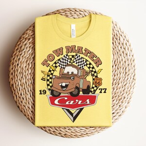 Puede incluir: Una camiseta de color amarillo claro con un gr&aacute;fico del personaje Tow Mater de la pel&iacute;cula Cars. El dise&ntilde;o incluye el texto Tow Mater, el n&uacute;mero 1977, una se&ntilde;al de la Ruta 66, banderas a cuadros, un trofeo y el logotipo rojo de Cars abajo.