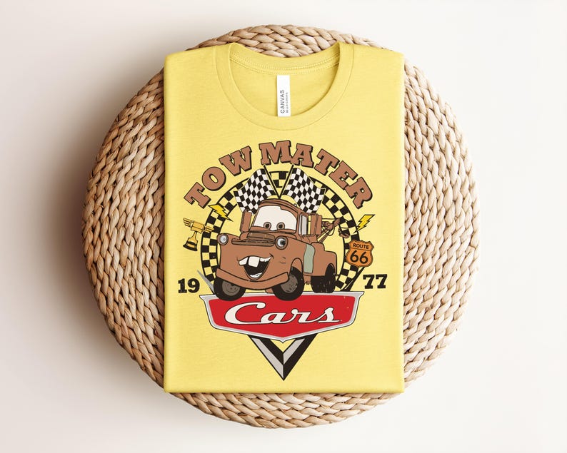 Puede incluir: Una camiseta de color amarillo claro con un gr&aacute;fico del personaje Tow Mater de la pel&iacute;cula Cars. El dise&ntilde;o incluye el texto Tow Mater, el n&uacute;mero 1977, una se&ntilde;al de la Ruta 66, banderas a cuadros, un trofeo y el logotipo rojo de Cars abajo.