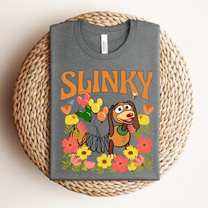 K&ouml;nnte beinhalten: Ein graues T-Shirt mit dem Wort "SLINKY" in orangefarbenen Buchstaben. Das Shirt zeigt ein Cartoon-Bild von Slinky Dog mit Luftballons, Blumen und einer Biene. Das T-Shirt ist auf einem gewebten, kreisf&ouml;rmigen Platzset gefaltet.