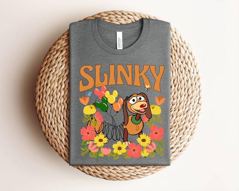 K&ouml;nnte beinhalten: Ein graues T-Shirt mit dem Wort "SLINKY" in orangefarbenen Buchstaben. Das Shirt zeigt ein Cartoon-Bild von Slinky Dog mit Luftballons, Blumen und einer Biene. Das T-Shirt ist auf einem gewebten, kreisf&ouml;rmigen Platzset gefaltet.
