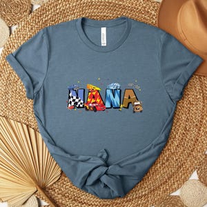 K&ouml;nnte beinhalten: Ein blaues T-Shirt mit dem Wort "NANA" in bunten Buchstaben, die jeweils mit Rennwagen und karierten Flaggen verziert sind. Das Shirt besteht aus einem weichen Material und hat kurze &Auml;rmel.