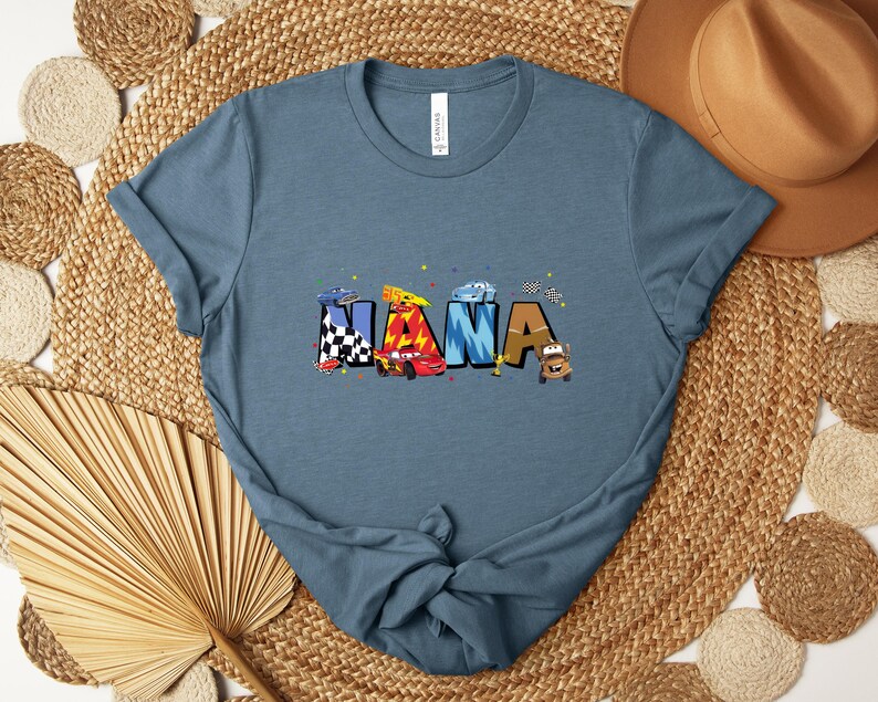 K&ouml;nnte beinhalten: Ein blaues T-Shirt mit dem Wort "NANA" in bunten Buchstaben, die jeweils mit Rennwagen und karierten Flaggen verziert sind. Das Shirt besteht aus einem weichen Material und hat kurze &Auml;rmel.