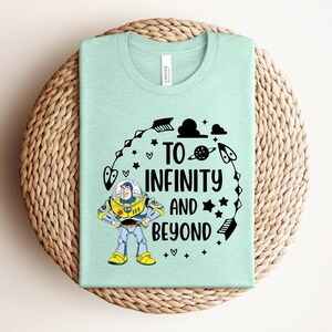 Peut inclure: Un t-shirt vert clair avec l'inscription "TO INFINITY AND BEYOND" en noir, ainsi qu'une image de dessin anim&eacute; de Buzz l'&Eacute;clair. Le design comprend des &eacute;toiles, des plan&egrave;tes et d'autres &eacute;l&eacute;ments sur le th&egrave;me de l'espace.