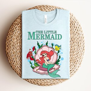 May include: A light blue t-shirt featuring a graphic of Ariel from The Little Mermaid sitting in a pink seashell, surrounded by green seaweed, a red crab, and a yellow fish. The text THE LITTLE MERMAID is printed in green above the illustration.