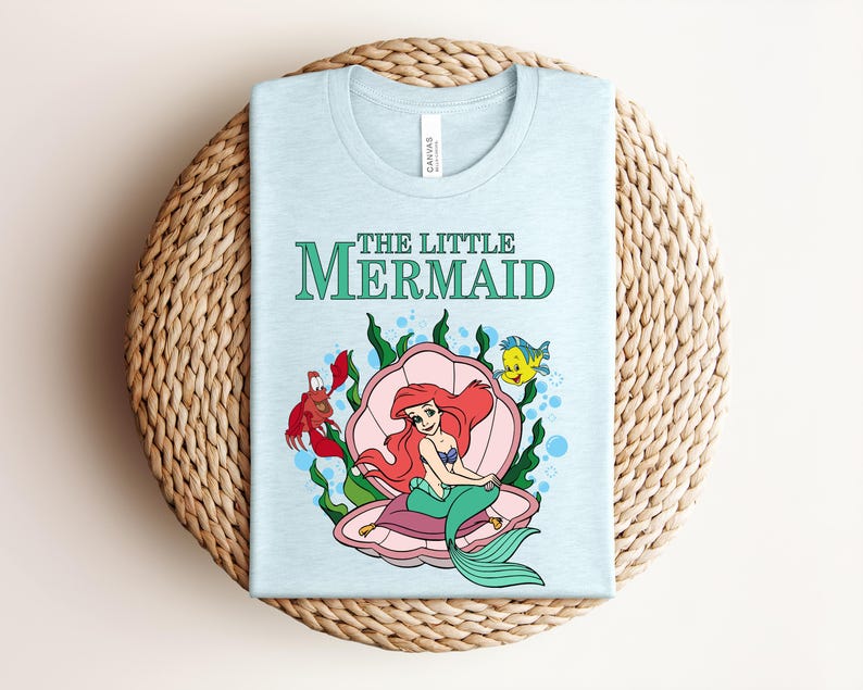 May include: A light blue t-shirt featuring a graphic of Ariel from The Little Mermaid sitting in a pink seashell, surrounded by green seaweed, a red crab, and a yellow fish. The text THE LITTLE MERMAID is printed in green above the illustration.