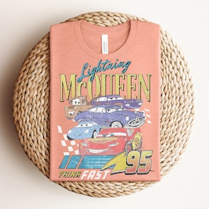 Puede incluir: Camiseta color melocot&oacute;n con un gr&aacute;fico de Rayo McQueen y otros personajes de Cars. El dise&ntilde;o incluye el texto "Lightning McQueen" y "Think Fast 95". La camiseta est&aacute; doblada sobre una superficie circular tejida.