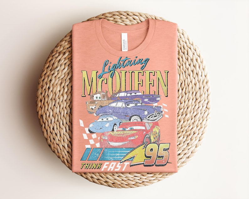 Puede incluir: Camiseta color melocot&oacute;n con un gr&aacute;fico de Rayo McQueen y otros personajes de Cars. El dise&ntilde;o incluye el texto "Lightning McQueen" y "Think Fast 95". La camiseta est&aacute; doblada sobre una superficie circular tejida.