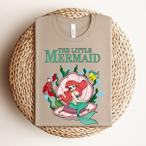 May include: A tan t-shirt featuring a graphic of The Little Mermaid with Ariel sitting in a pink seashell, surrounded by green seaweed, a red crab, and a yellow fish. The text THE LITTLE MERMAID is printed in teal above the character illustration.
