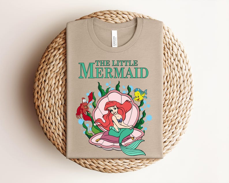 May include: A tan t-shirt featuring a graphic of The Little Mermaid with Ariel sitting in a pink seashell, surrounded by green seaweed, a red crab, and a yellow fish. The text THE LITTLE MERMAID is printed in teal above the character illustration.