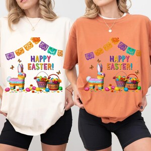 K&ouml;nnte beinhalten: Zwei T-Shirts, eines cremefarben und eines orangefarben, zeigen ein farbenfrohes Osterdesign. Das Design umfasst einen Pi&ntilde;ata-Hasen, Eierk&ouml;rbe, Schmetterlinge und den Text "HAPPY EASTER!" &uuml;ber der Bildsprache, mit dekorativen Bannern.