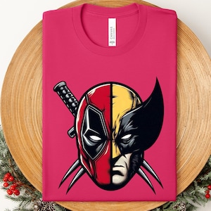 May include: A folded magenta t-shirt featuring a split face design. One half is Deadpool's mask, the other Wolverine's face. The design includes Deadpool's katana and Wolverine's claws. The shirt is on a wooden surface.